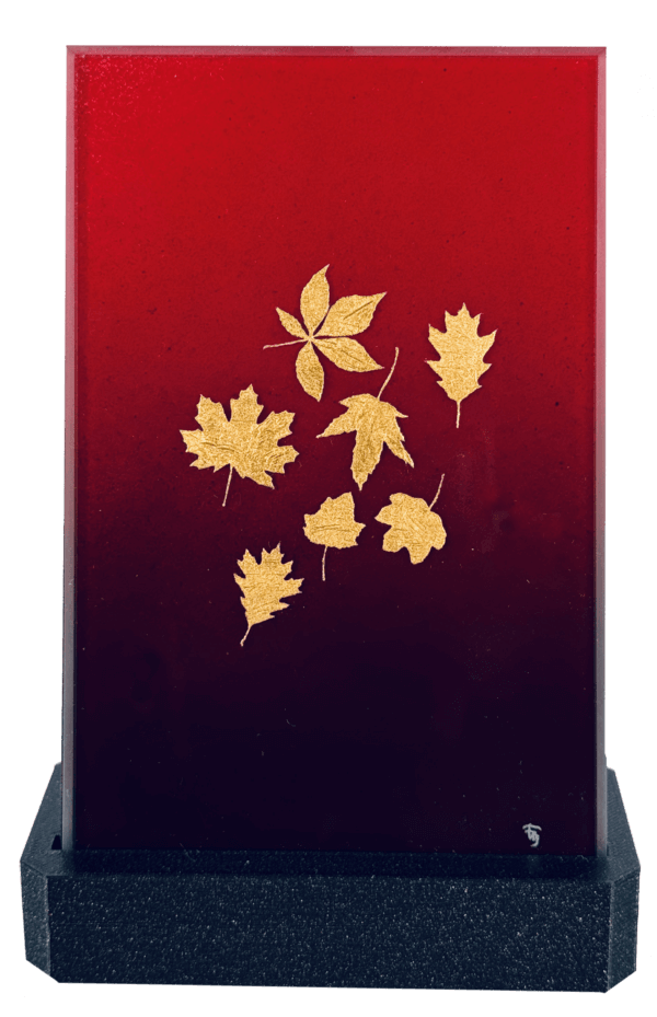Rote Glasplatte mit goldenen Blattern, stehend auf schwarzem Sockel, herbstliche Eleganz und Dekoration.