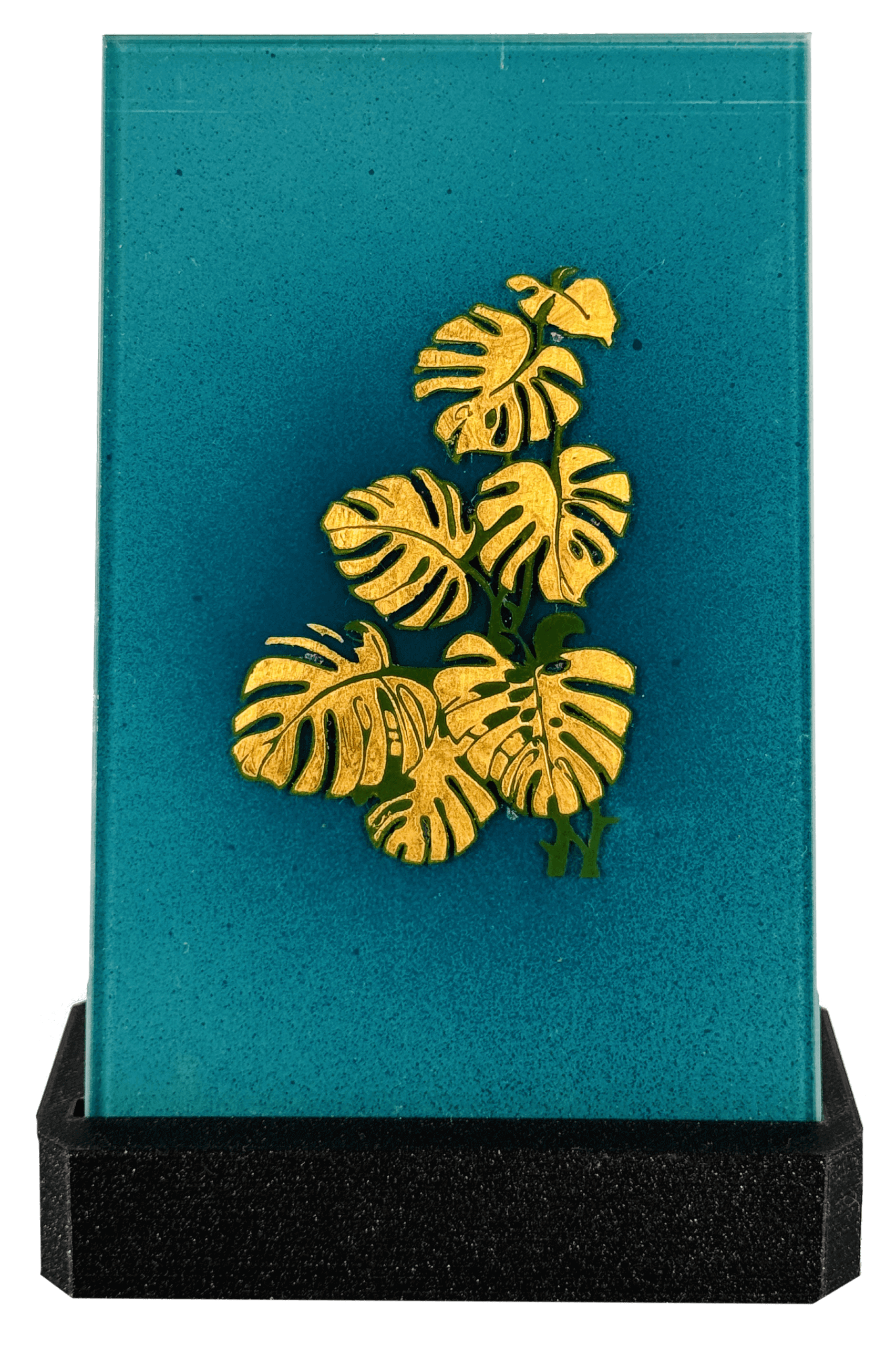 Goldene Monstera-Blätter auf türkisfarbenem Glasblock mit schwarzem Sockel, elegantes Deko-Element.