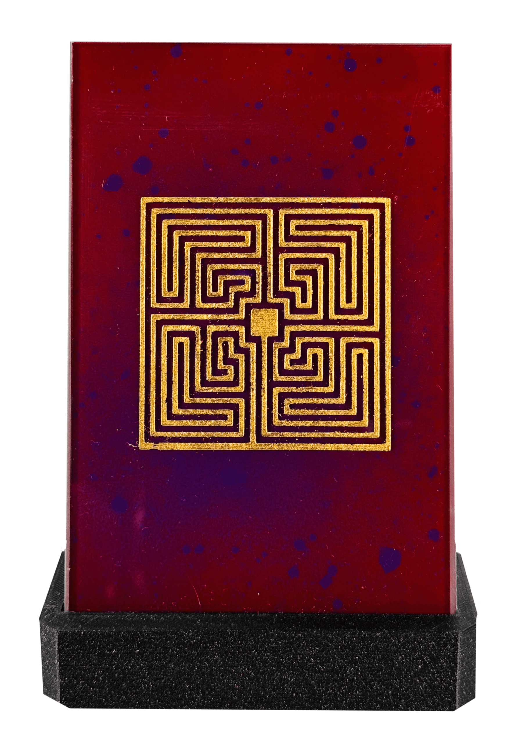 Rotes Kunstwerk mit goldenem Labyrinth-Design auf schwarzem Sockel, verziert mit blauen Sprenkeln.